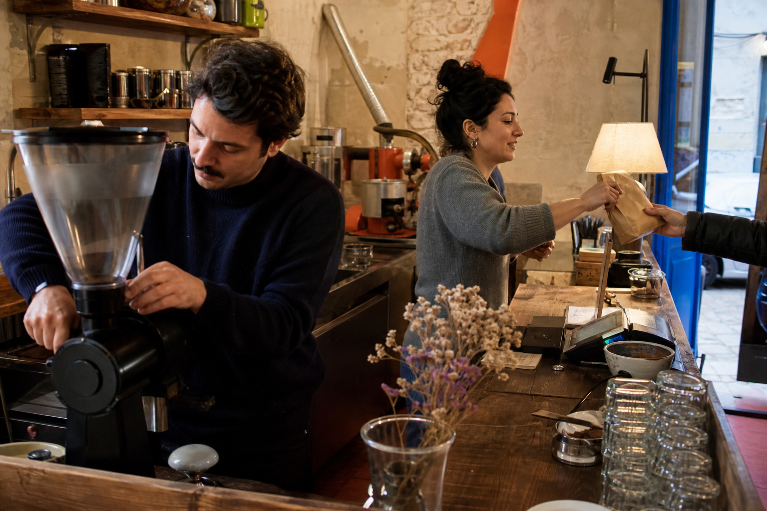 Due persone al lavoro dietro il bancone: uno macina il caffè, l’altra consegna un sacchetto